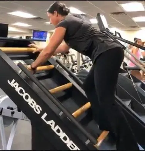 Woman exercising on a Jacobs Ladder machine in a gym, holding onto the rungs, dark clothing.