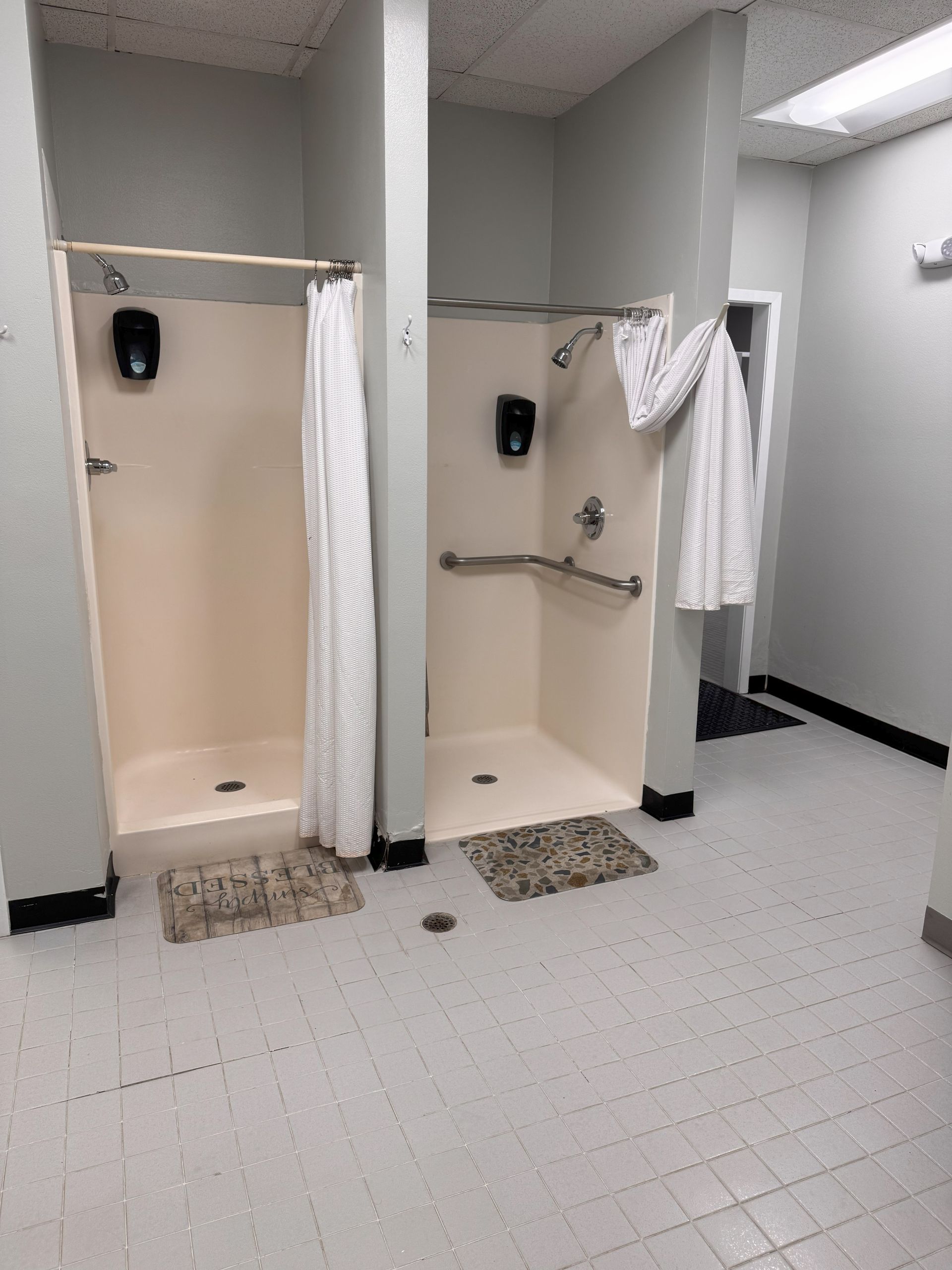 Two shower stalls in a bathroom; beige walls, white curtains, grab bar, gray floor mats, black soap dispensers.
