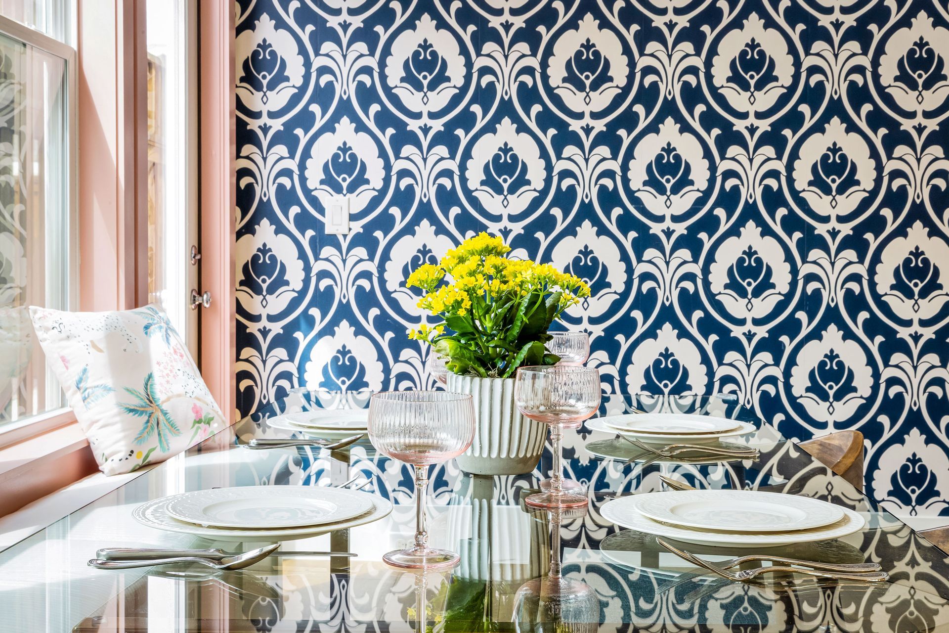 Elegant dining nook with blue-and-white patterned wallpaper, glass table, and yellow flowers by a window