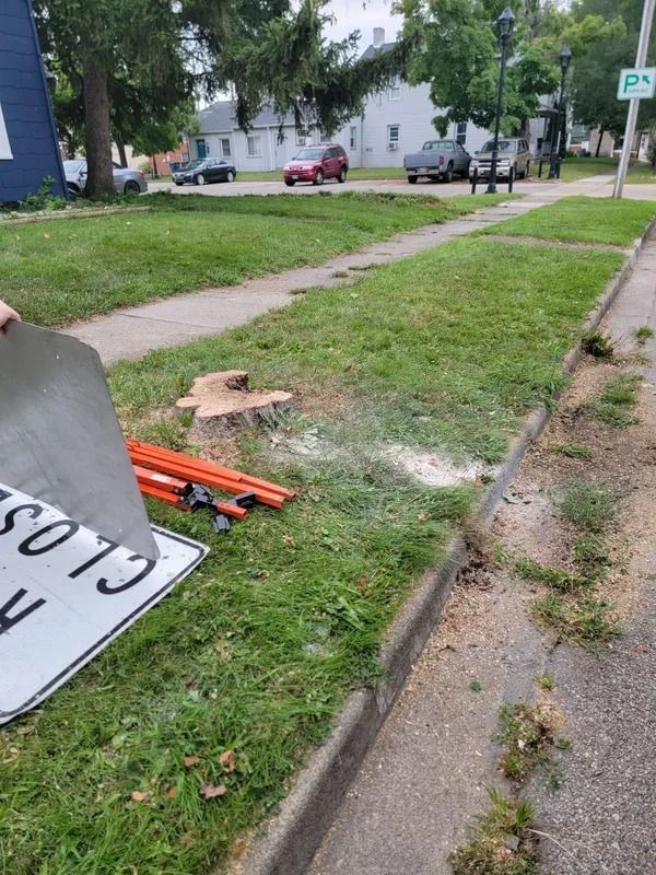Tree stump on a grassy median with road closure sign and construction equipment near a sidewalk and street.