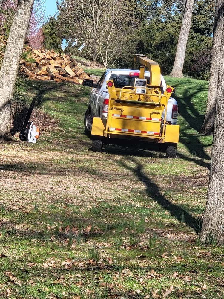 A yellow wood chipper attached to a truck, parked on a grassy slope next to a pile of wood.