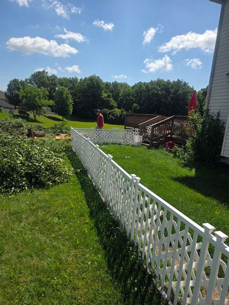 White lattice fence in a grassy yard, with a person standing by a deck and trees in the background under a blue sky.