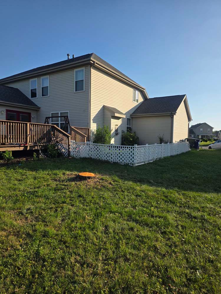 Back of a two-story beige house with a wooden deck, white fence, and green lawn on a sunny day.