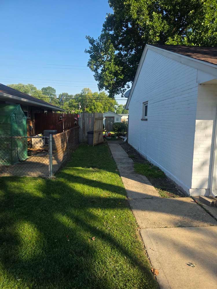 A narrow pathway beside a white building and fence, with green grass and trees in the background under a blue sky.