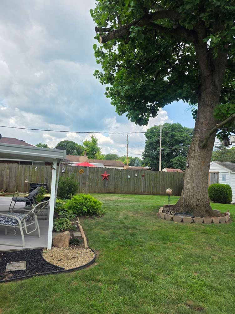 Backyard with a large tree, green lawn, a fence, and a patio with chairs under a canopy.