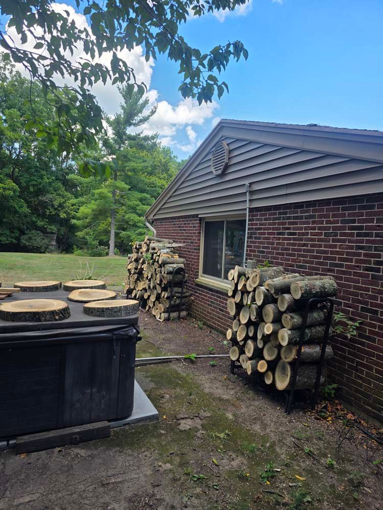 Stacks of firewood next to a brick house with a window, near a hot tub. Blue sky and trees in the background.