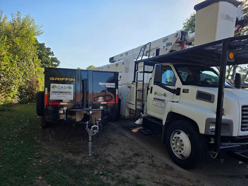 Tree service truck and trailer parked on grass. The trailer is black and the truck is white.