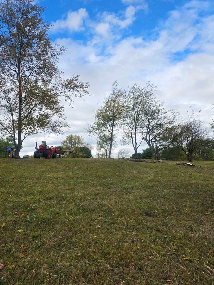 A red tractor on a grassy hill under a partly cloudy sky, trees scattered around the tractor.