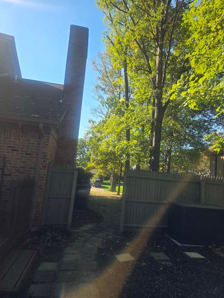 Brick house and chimney with open wooden gate leading to a path under trees in a sunny outdoor setting.