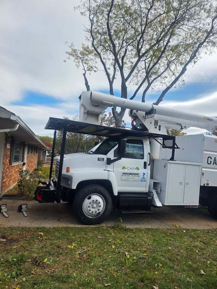 White tree service truck with extended lift arm parked in front of a house, cutting tree branches.