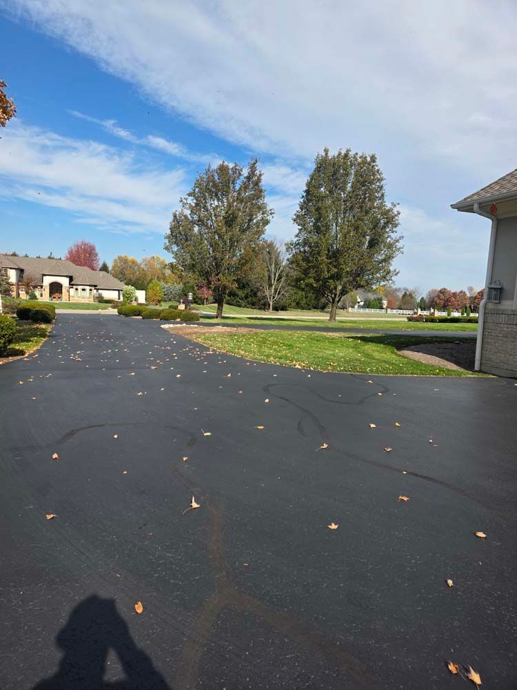 Asphalt driveway with scattered leaves leading to a grassy area, trees, and houses under a partly cloudy sky.