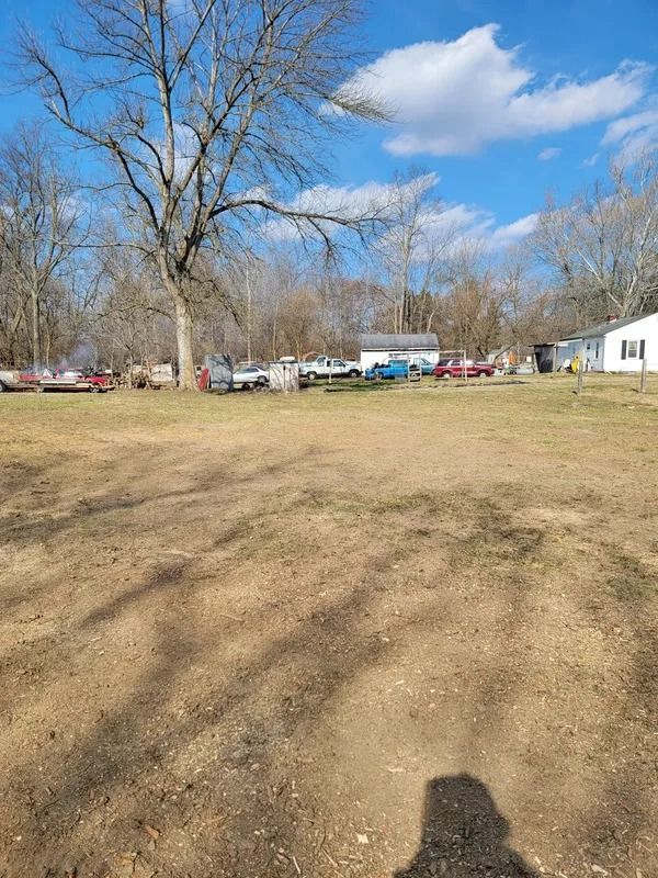 Brown field with several parked vehicles in the distance on a sunny day. Bare trees and a small white building are also visible.