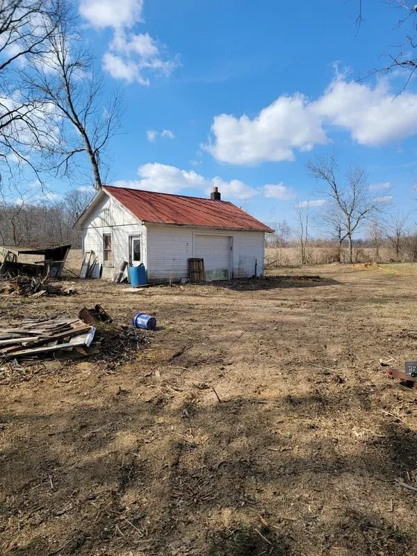 Old white house with red roof on a brown field under a blue sky. Bare trees surround it.