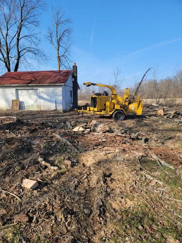 A yellow wood chipper beside a small, white building in a muddy field under a blue sky.
