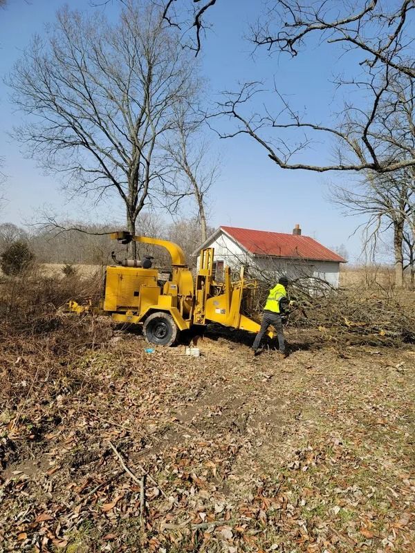 Yellow wood chipper with worker chipping branches next to a small building under a blue sky.