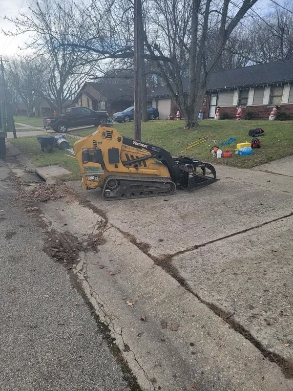 Yellow skid steer with cutting attachment parked on street near houses.