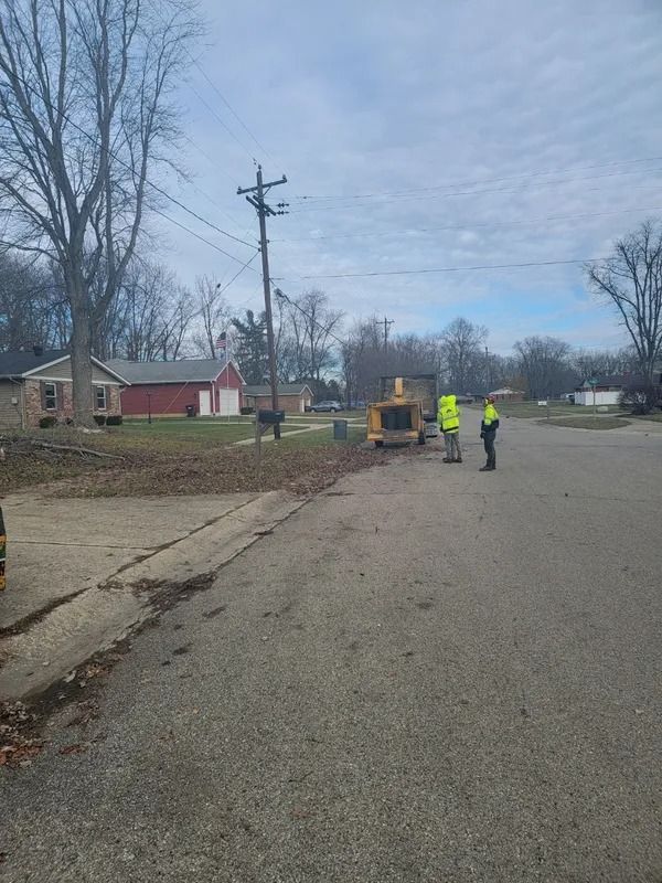 Two workers in safety vests operating equipment on a street, likely for tree trimming or utility work.