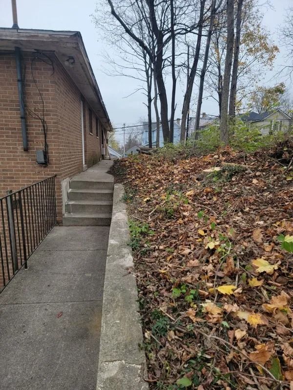 Concrete walkway with steps and railing next to a brick building and a leaf-covered hill.