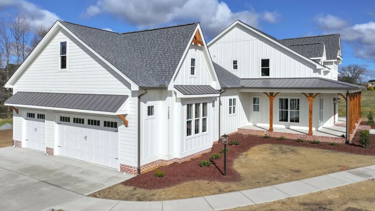 A white farmhouse-style home with a grey roof, attached garage, and a wrap-around porch, set against a blue sky.