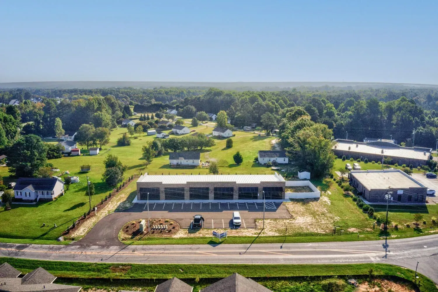 Aerial view of a modern, multi-unit commercial strip mall with a parking lot along a road in a green, wooded area.