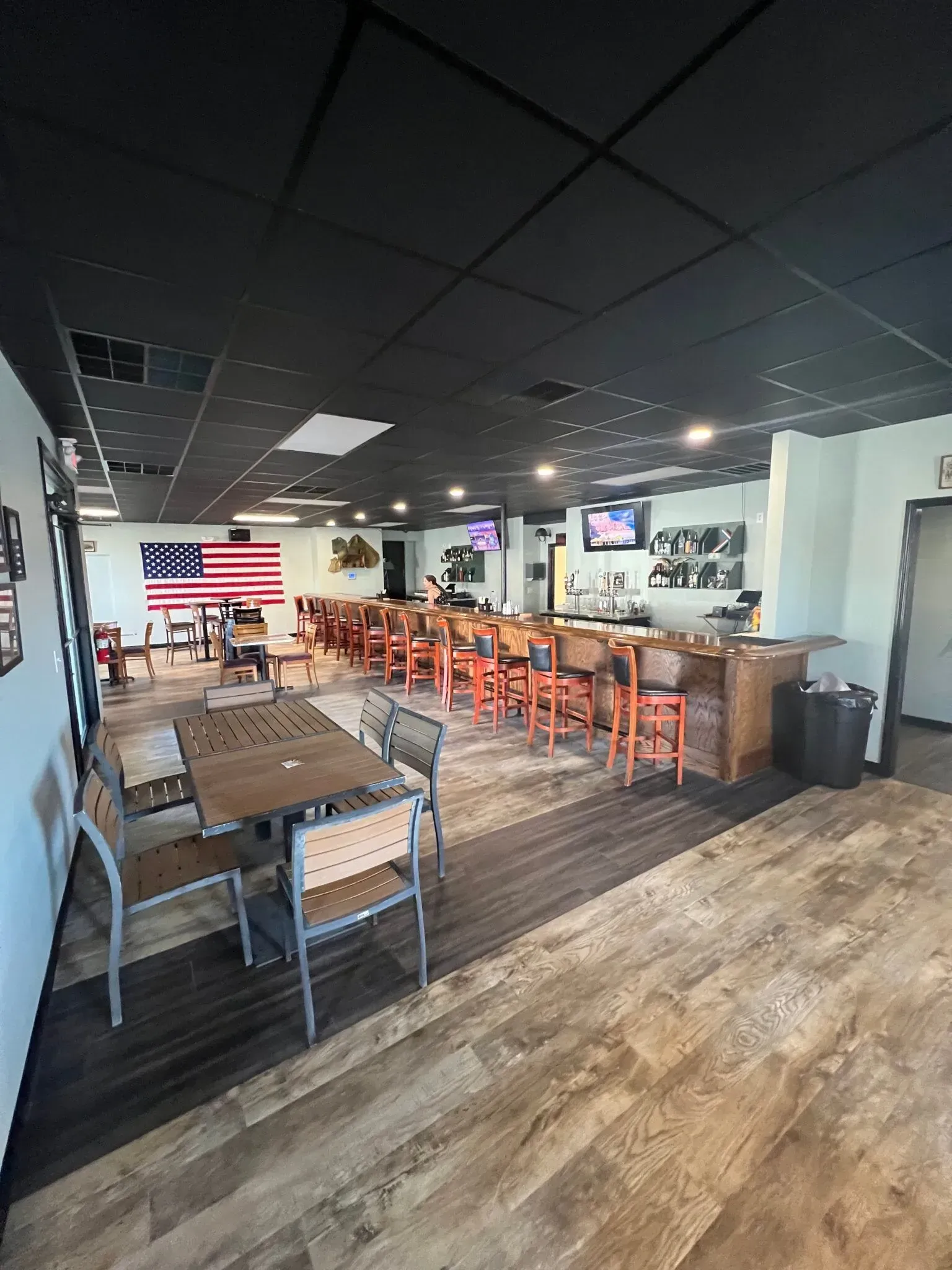 A restaurant interior featuring rows of tables, a long bar with stools, and a large American flag on the back wall.