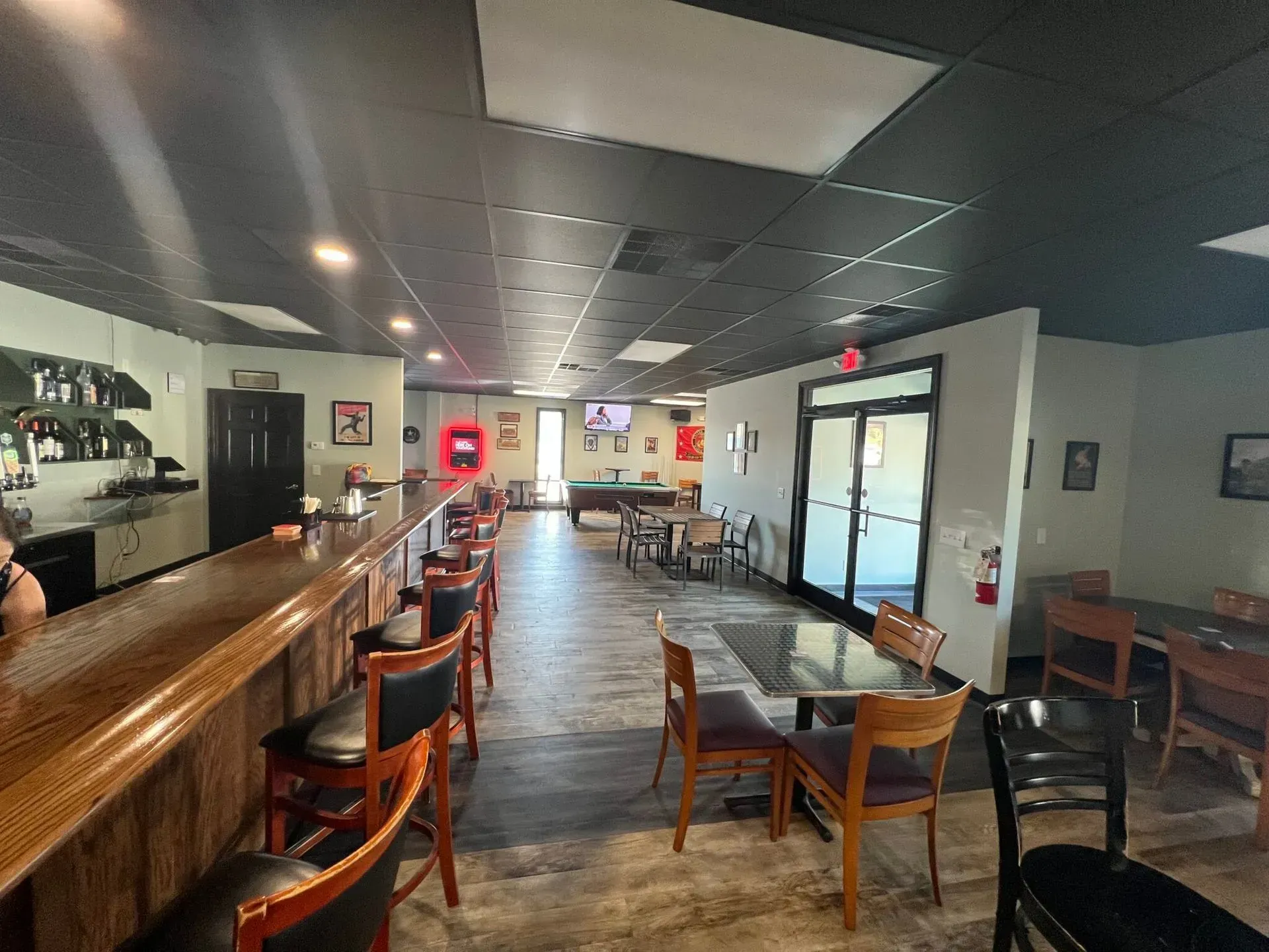 An interior view of a bar with a wooden counter, high chairs, tables with chairs, and a pool table in the distance.