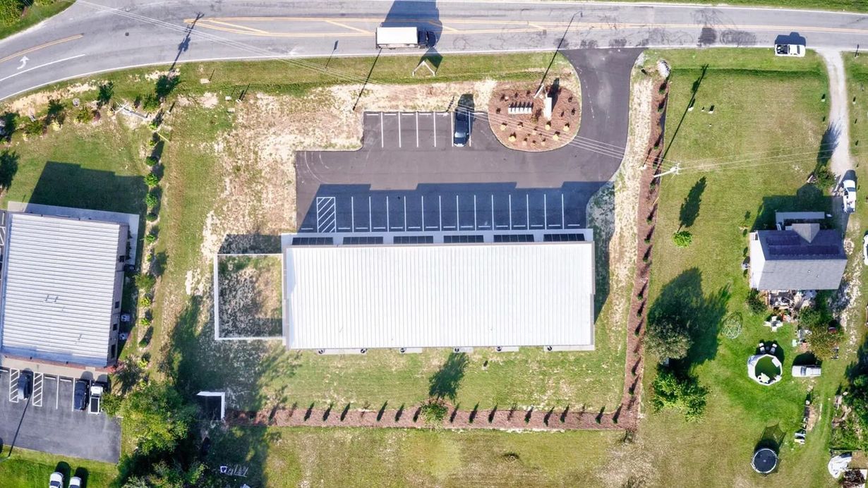 Aerial view of a commercial building with a long white roof, parking lot, and adjacent lawn in a rural setting.
