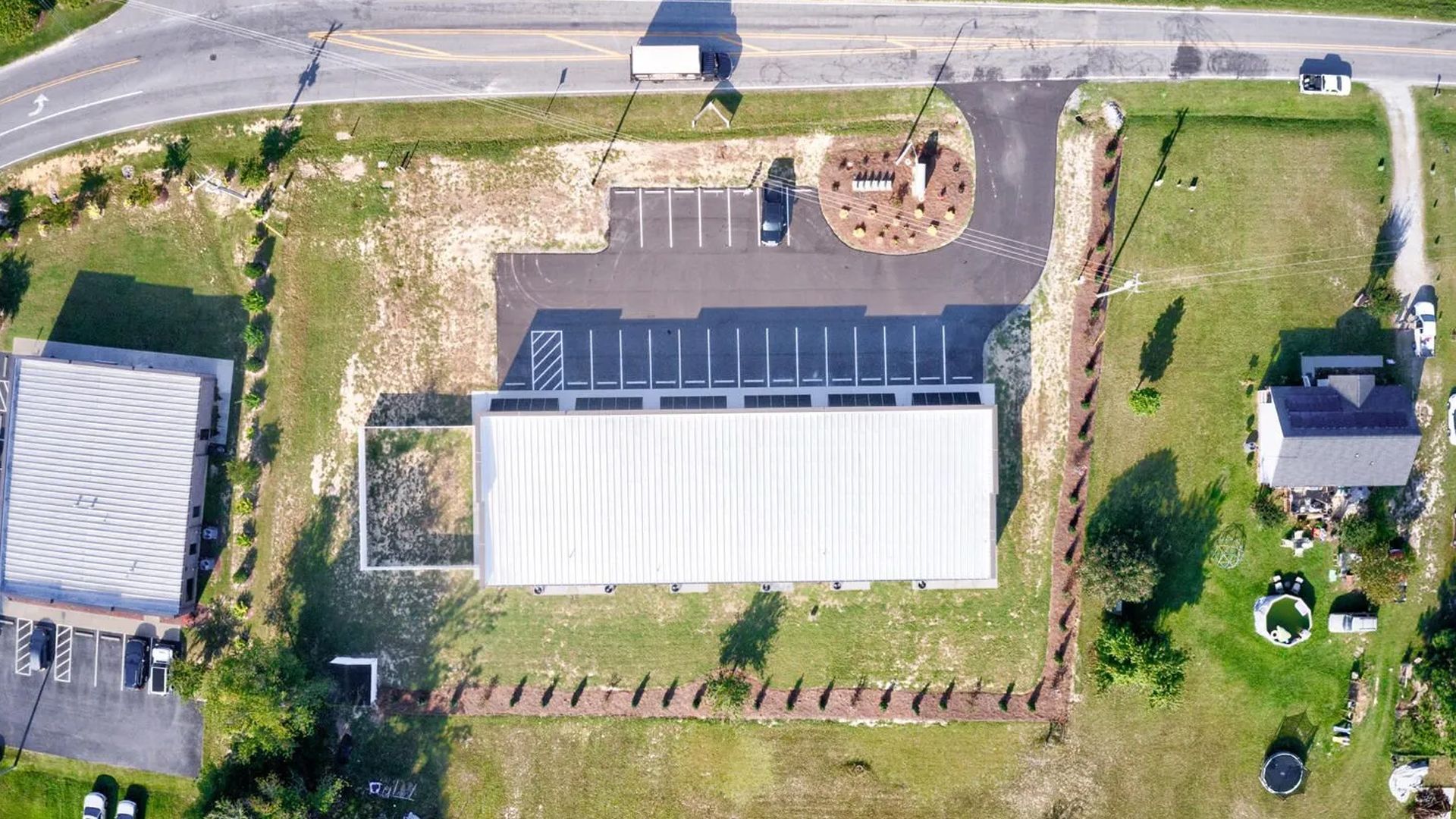 Aerial view of a commercial building with a long white roof, parking lot, and adjacent lawn in a rural setting.