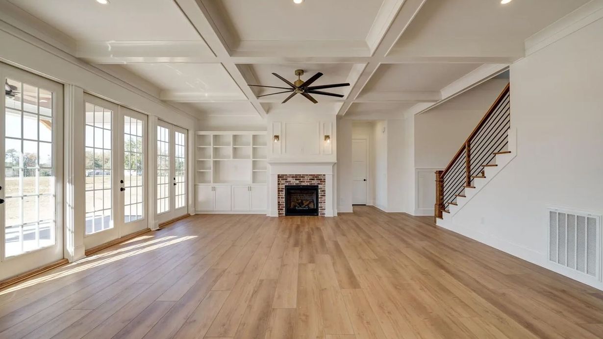 A spacious, light-filled living room featuring white coffered ceilings, light wood floors, a fireplace, and a staircase.