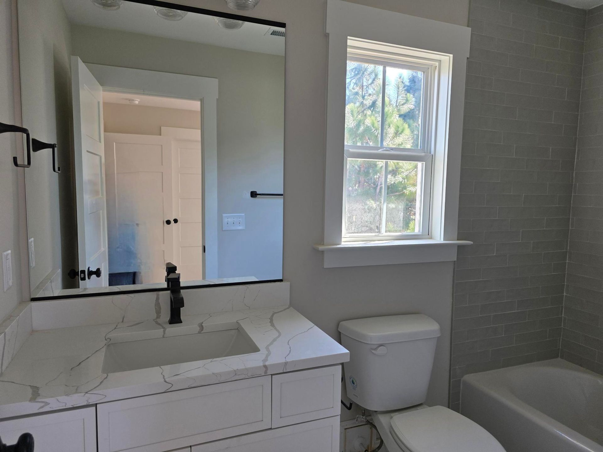 Modern bathroom with white vanity, mirror, toilet, bathtub, and a window.