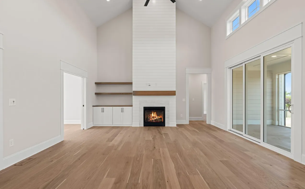 Bright empty living room with hardwood floors, tall white fireplace, built-in shelves, and sliding glass doors