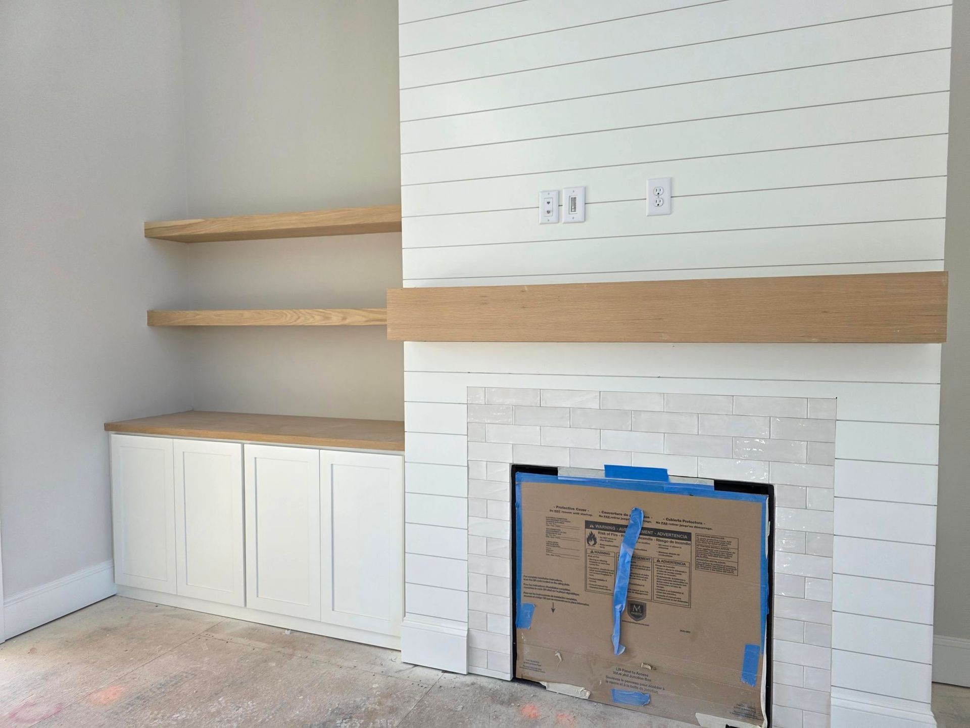 Living room fireplace with white tiled surround, wood mantel, floating shelves, and white cabinets