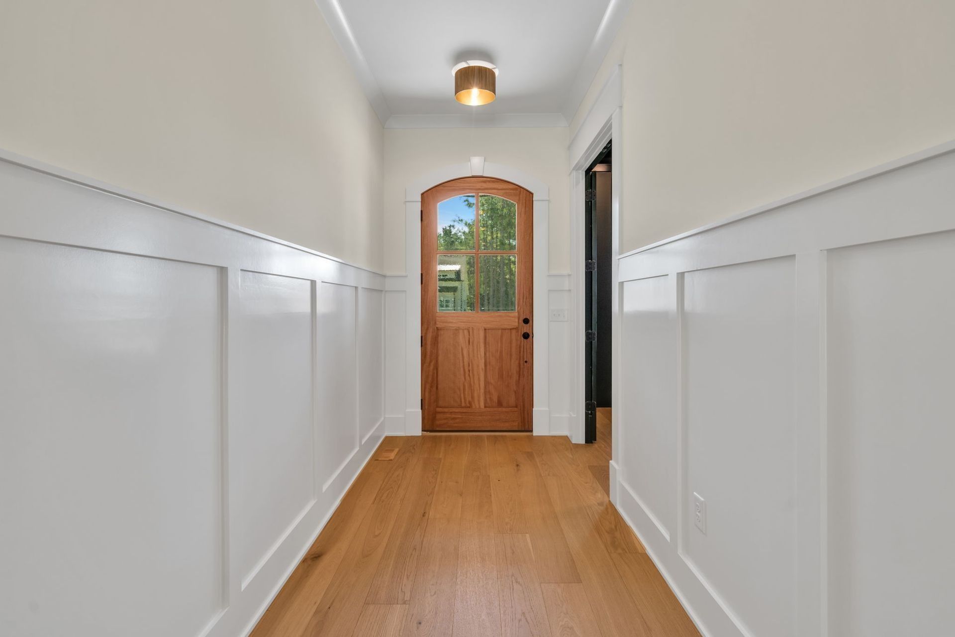 Bright hallway with white wainscoting, wood floor, and a wooden front door at the end.