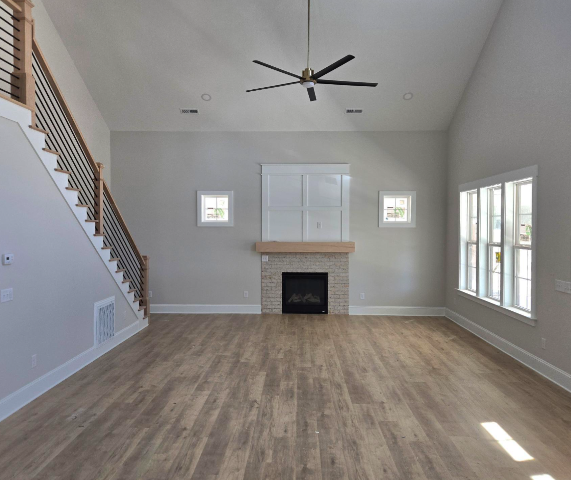 Empty living room with wood floor, fireplace, ceiling fan, and large windows.
