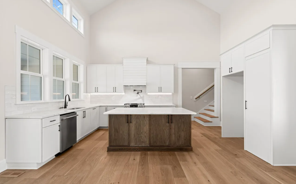 Bright modern white kitchen with island, wood floor, and staircase in the background