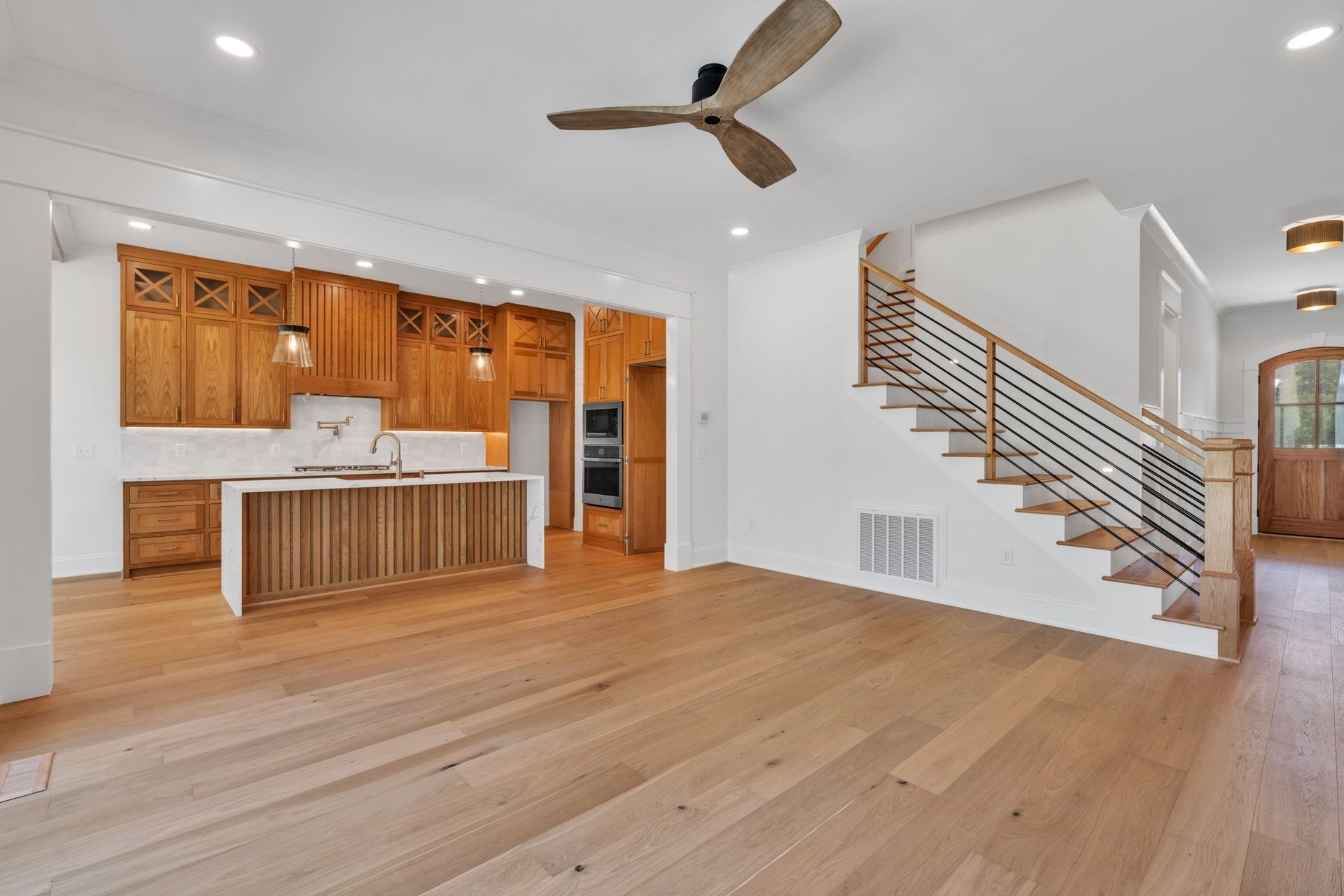 Open living room with wood floors, kitchen island, and staircase with metal railing under ceiling fan
