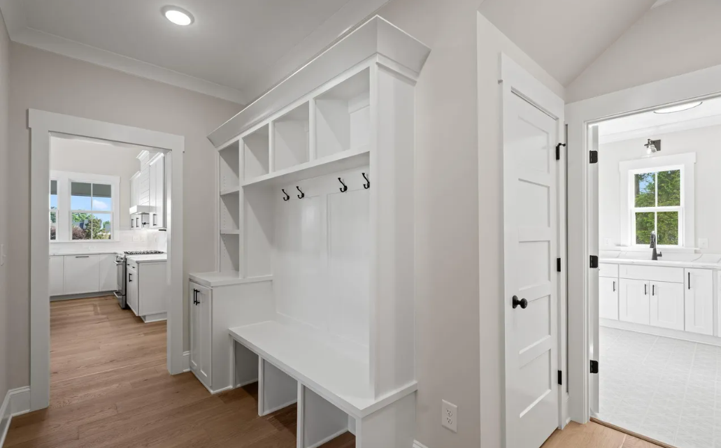 Bright mudroom with white built-in bench and hooks, leading to a white bathroom with a window.