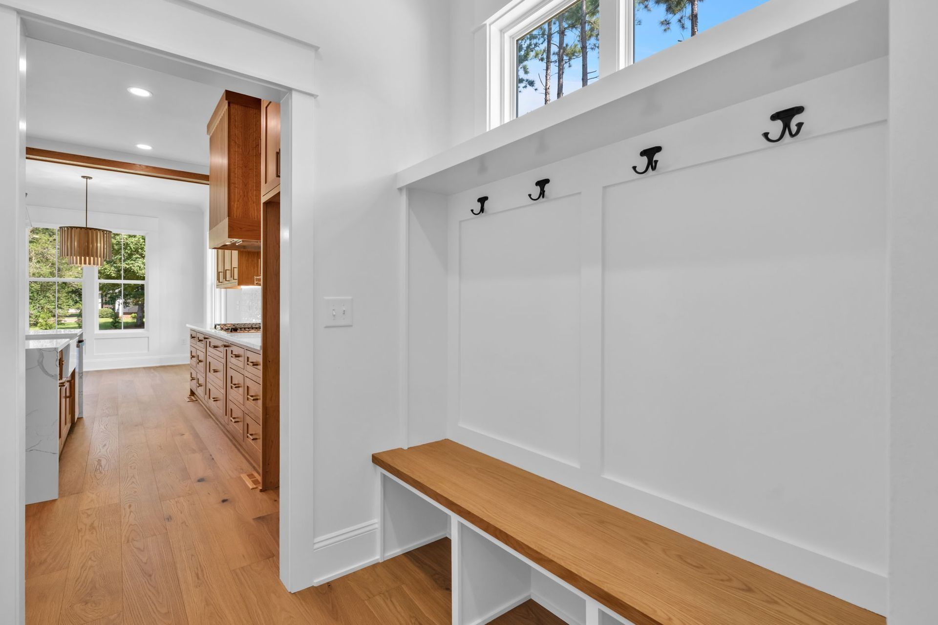 White mudroom with bench and hooks, opening to a bright kitchen with wood floors and cabinets