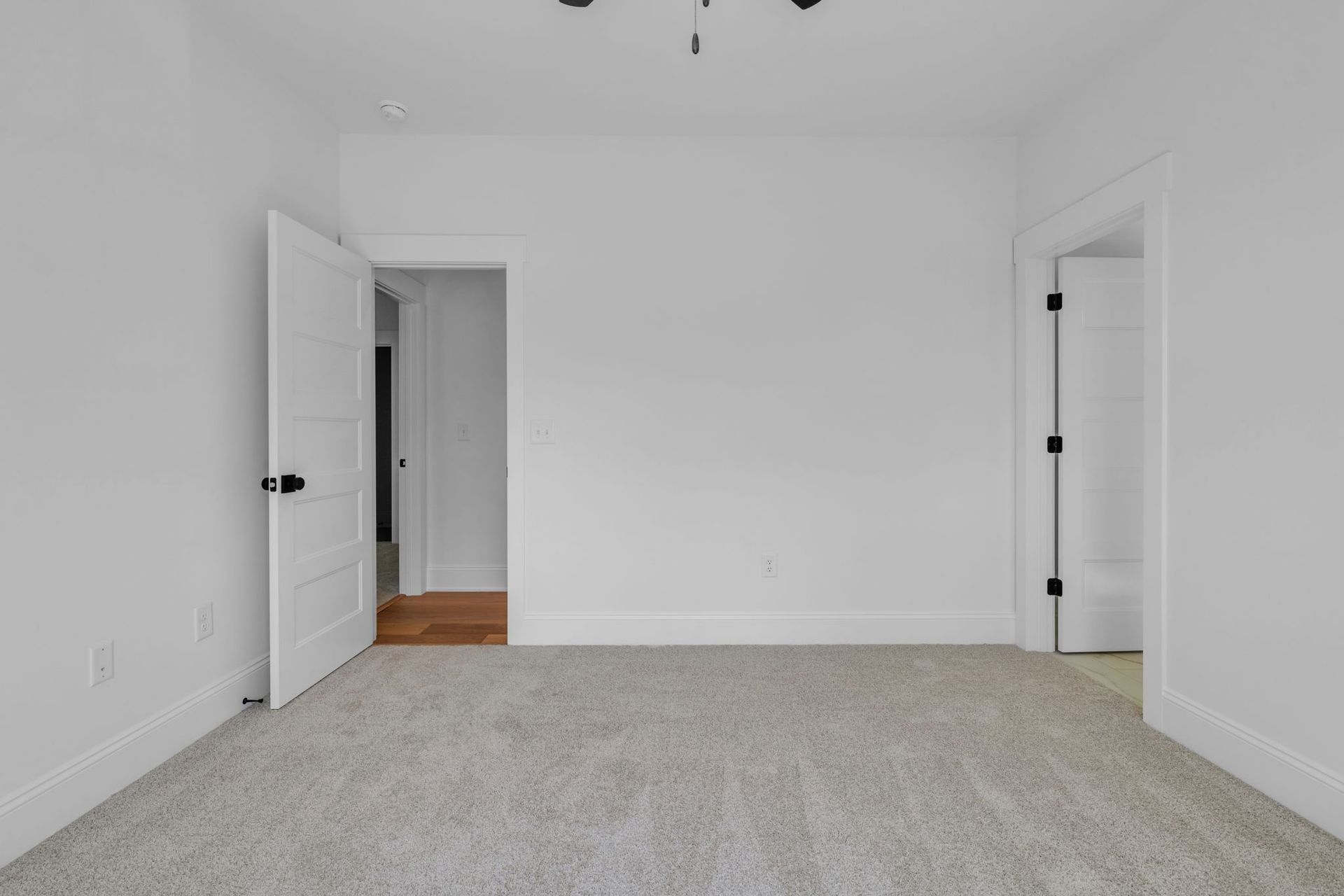Empty white bedroom with open doors, beige carpet, and wood floor visible in the hallway