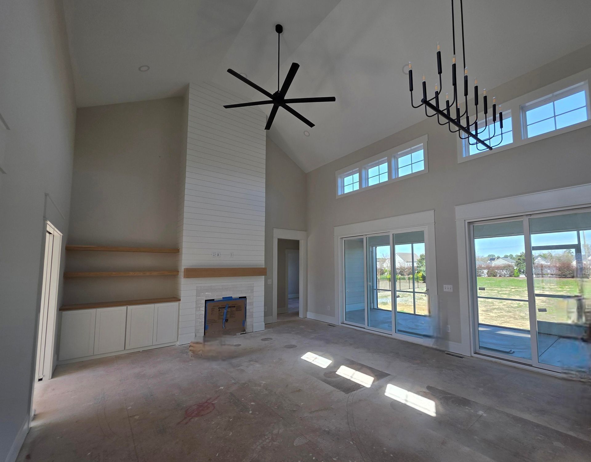 Bright empty living room with high ceilings, fireplace, chandeliers, and large windows overlooking a yard