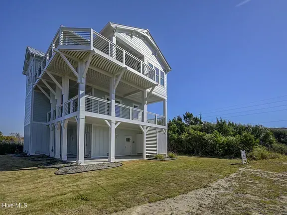 White multi-story coastal house with balconies and stairs on a grassy lot under a clear blue sky