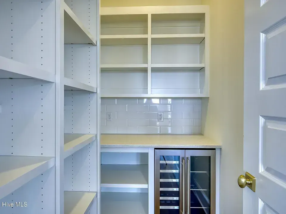 Empty white pantry with built-in shelves, countertop, and under-cabinet wine cooler; door open.