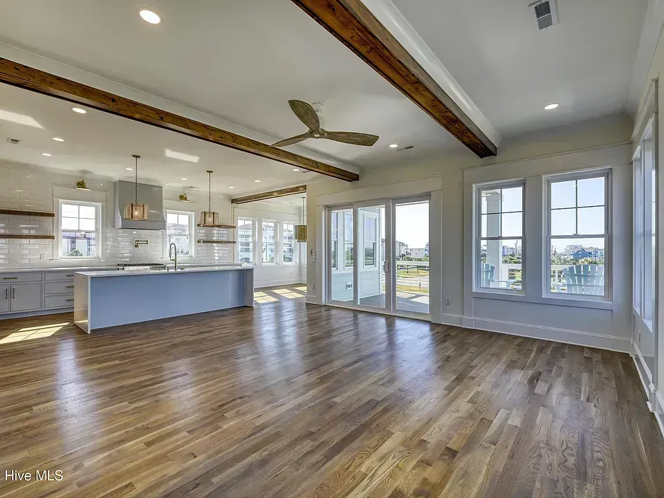 Bright open-plan living room with hardwood floors, exposed beams, ceiling fan, and a kitchen island.