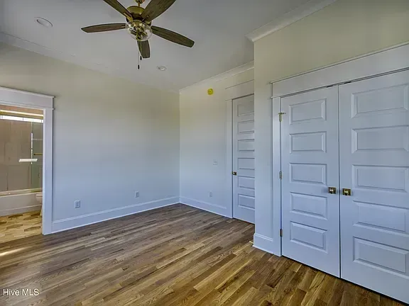Empty bedroom with white walls, wood floors, ceiling fan, and closet doors