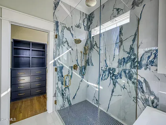 Marble-tiled walk-in shower with glass panel and built-in shelf beside an open doorway to a dark dresser room