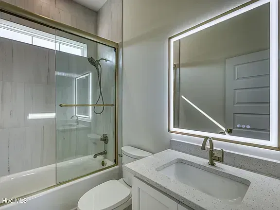 Bright modern bathroom with a large backlit mirror, white vanity, and shower-tub combo with glass enclosure.