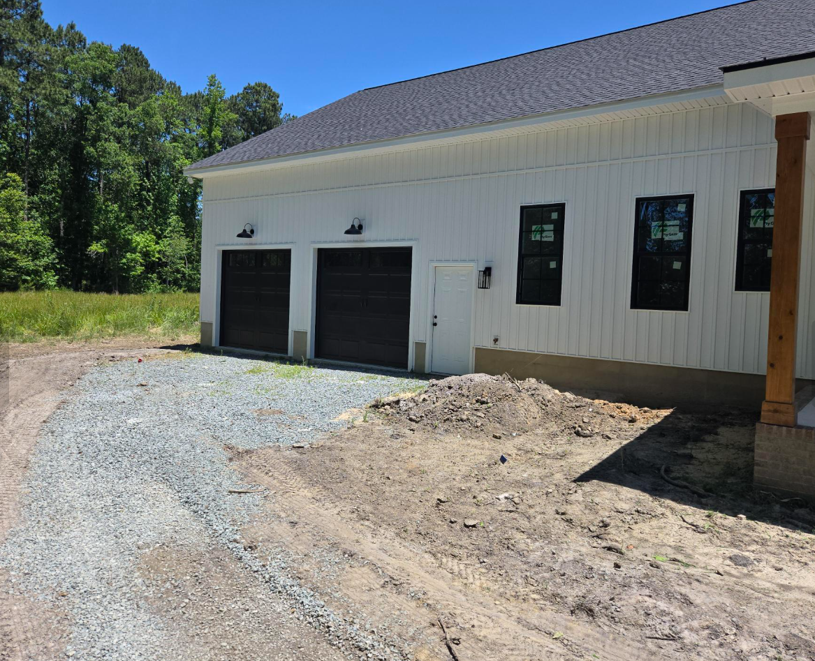 White building with two black garage doors, a gravel driveway, and a dirt area beside a wooded lot