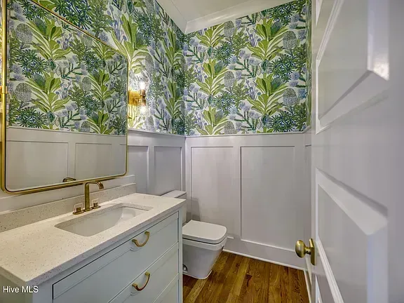 Bright bathroom with tropical wallpaper, white wainscoting, vanity, mirror, and toilet.