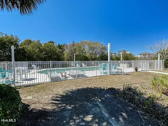 Outdoor pool area with white fence, turquoise water, lounge chairs, and trees under a clear blue sky