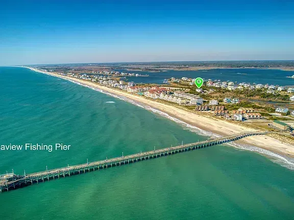 Aerial view of a sandy beach with turquoise water, a long fishing pier, and coastal homes under a clear blue sky